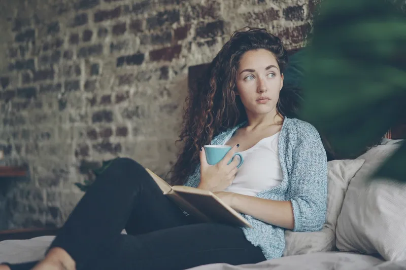 A woman resting on a bed with a book and a mug, looking out of the frame.