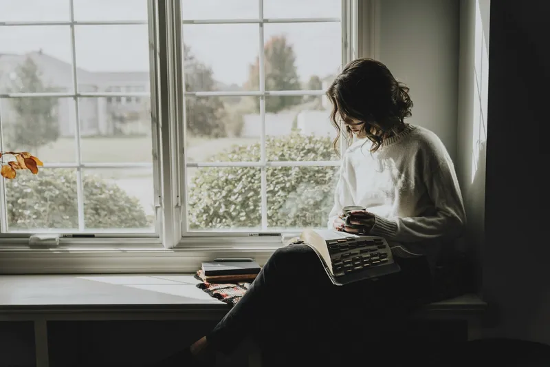 A woman sitting in a window seat with a book and a mug.